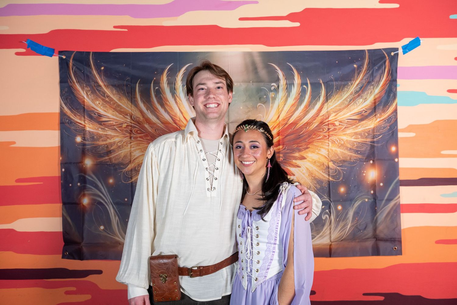 Couples pose for photos in front of glowing wing mural with sunset background.