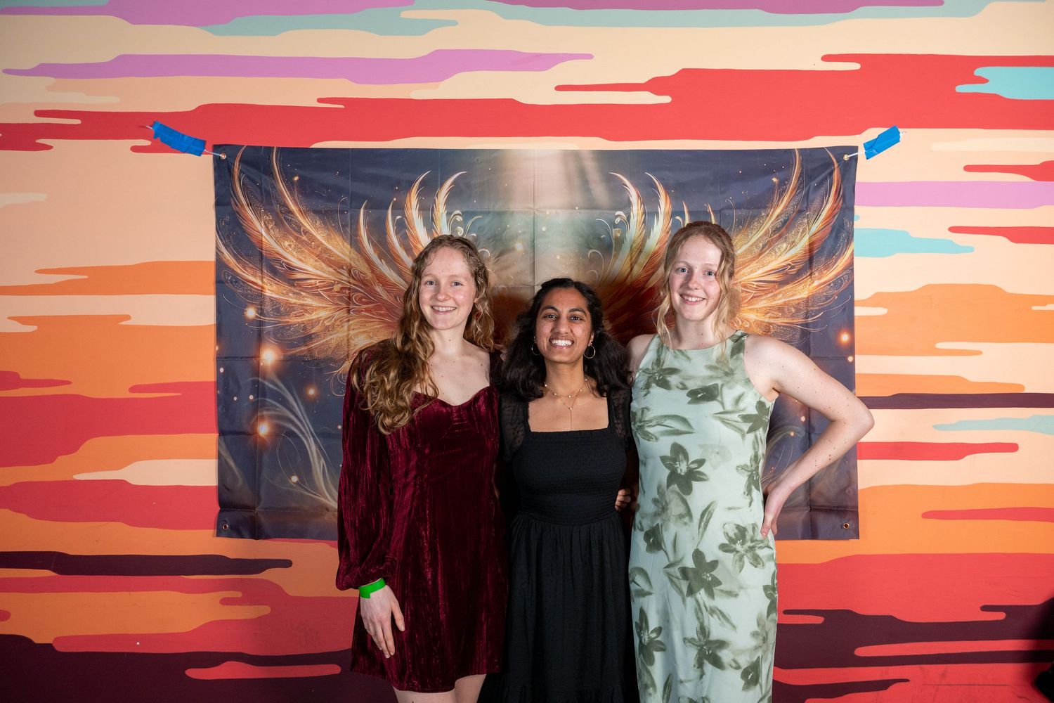 Friends gather for photo in front of artistic wing mural with warm sunset tones.