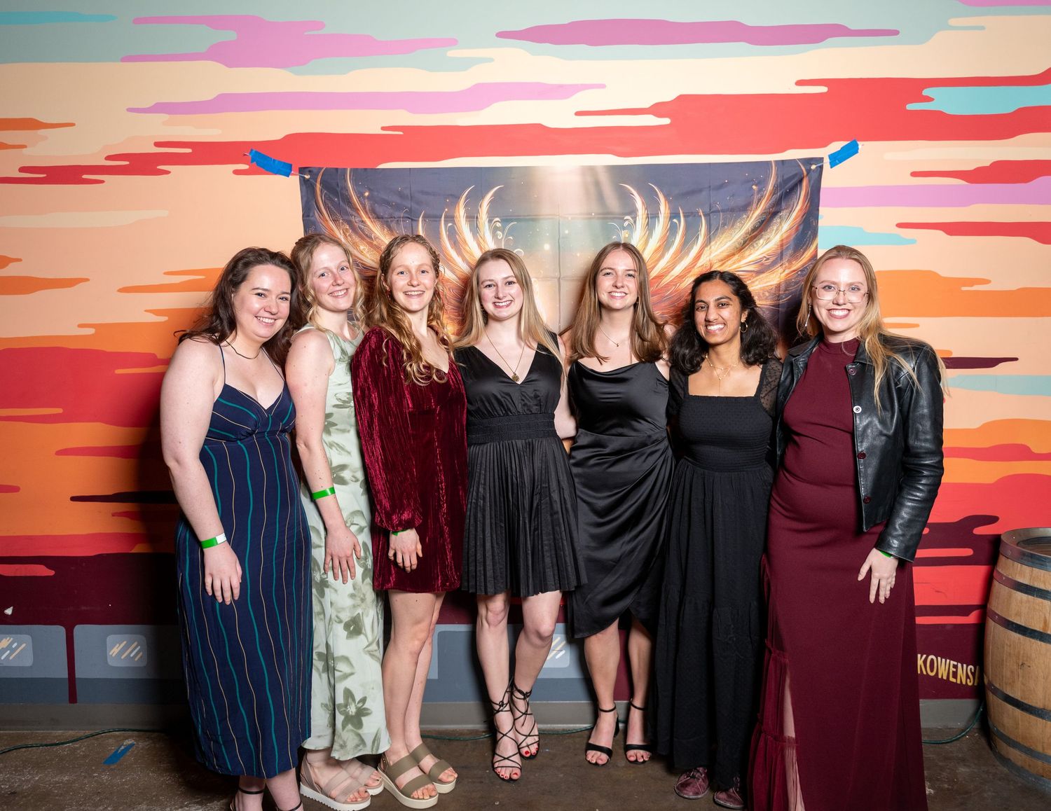 Group wearing dark formal attire poses together against wing-themed backdrop.