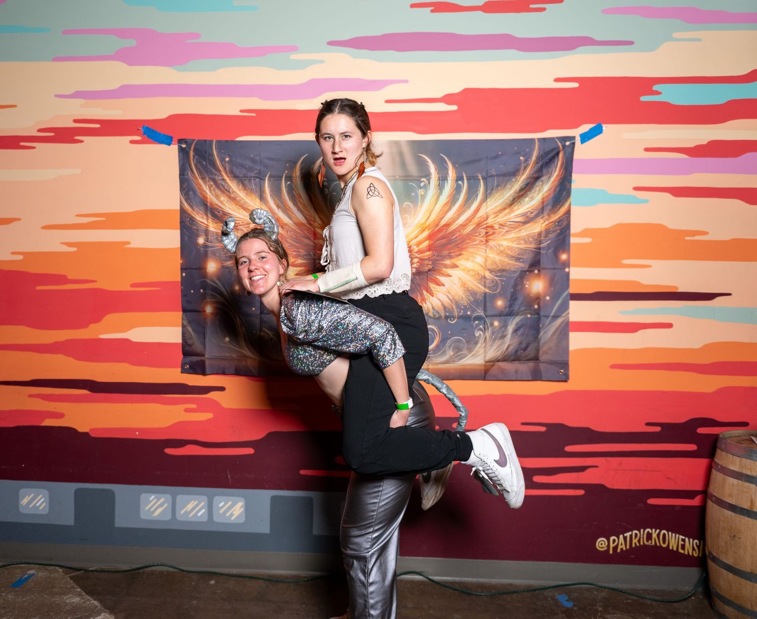 A person poses playfully in front of a colorful sunset-themed wall mural with golden wings.