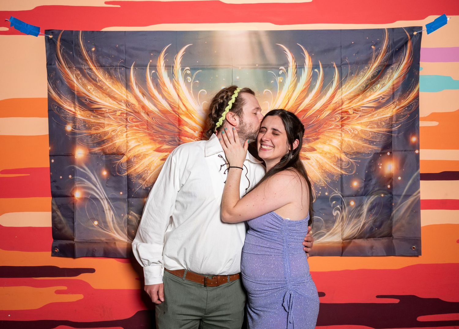 Friends pose playfully against colorful sunset backdrop with glowing wing design.