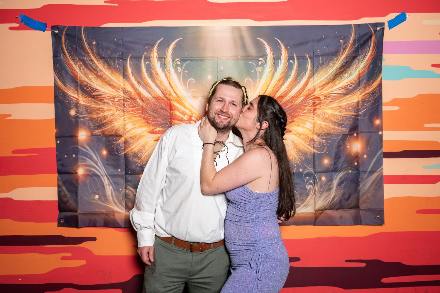 Couples pose playfully against a dramatic sunset backdrop with illuminated angel wings.