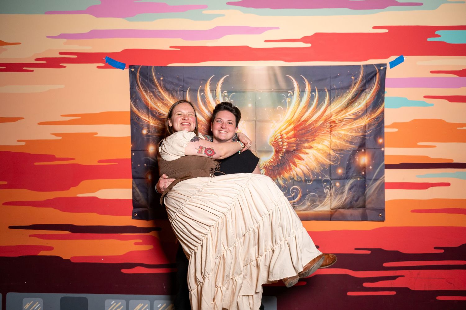 Party attendees strike fun poses against sunset-themed photo backdrop with golden wings.