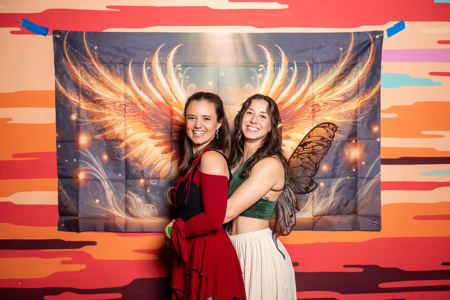 Two people in formal attire standing before a sunset-themed mural with golden wings.