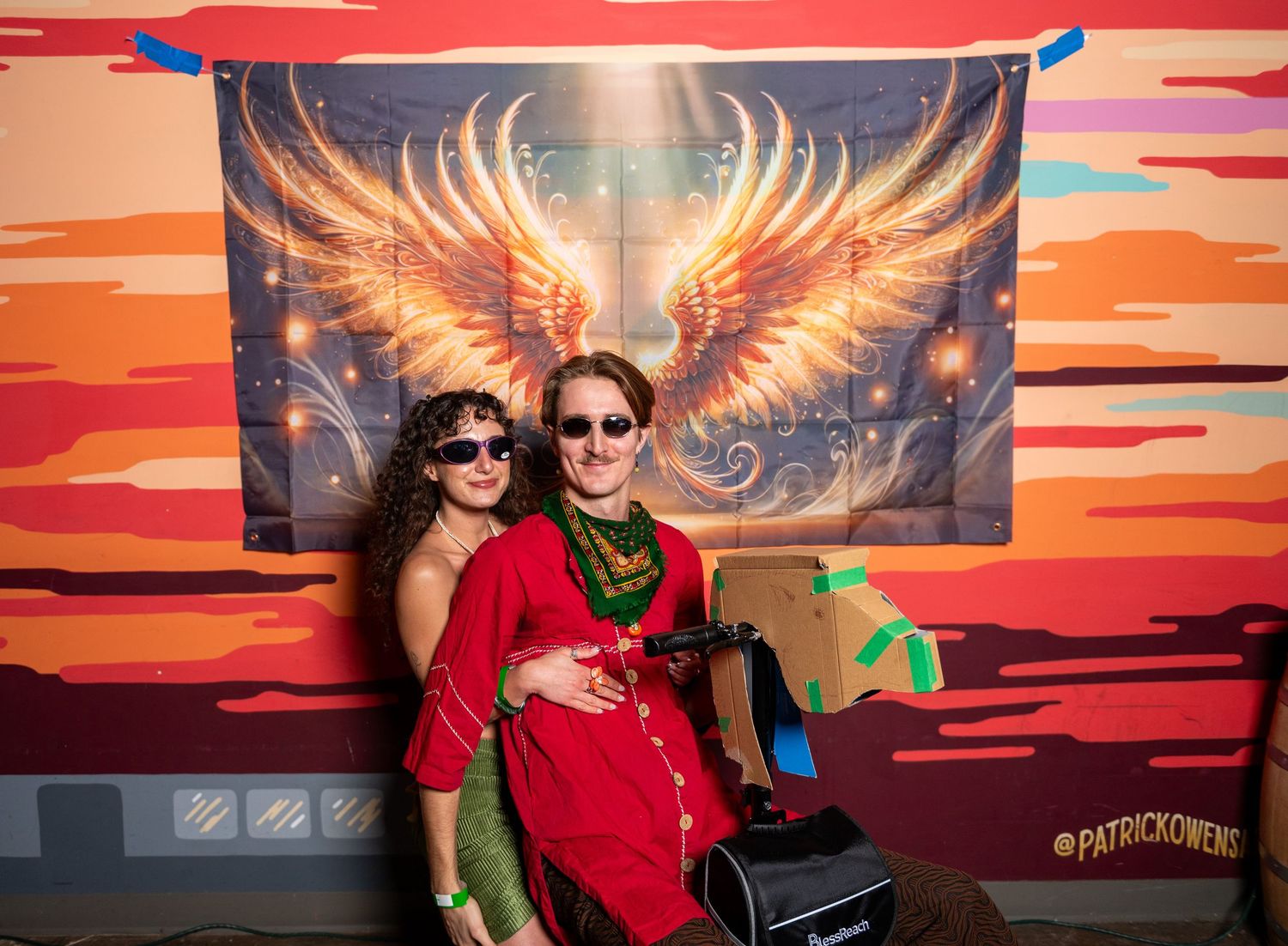 Friends gather for photos against a dramatic angel wing mural with sunset-colored background.