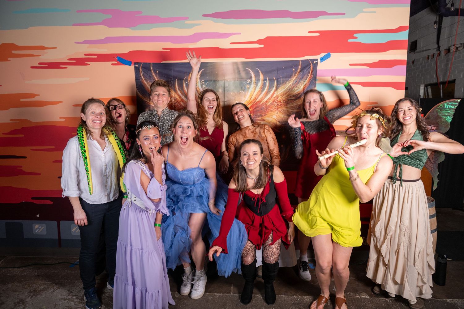 Group poses together in front of vibrant sunset backdrop with painted golden wings at a party event.