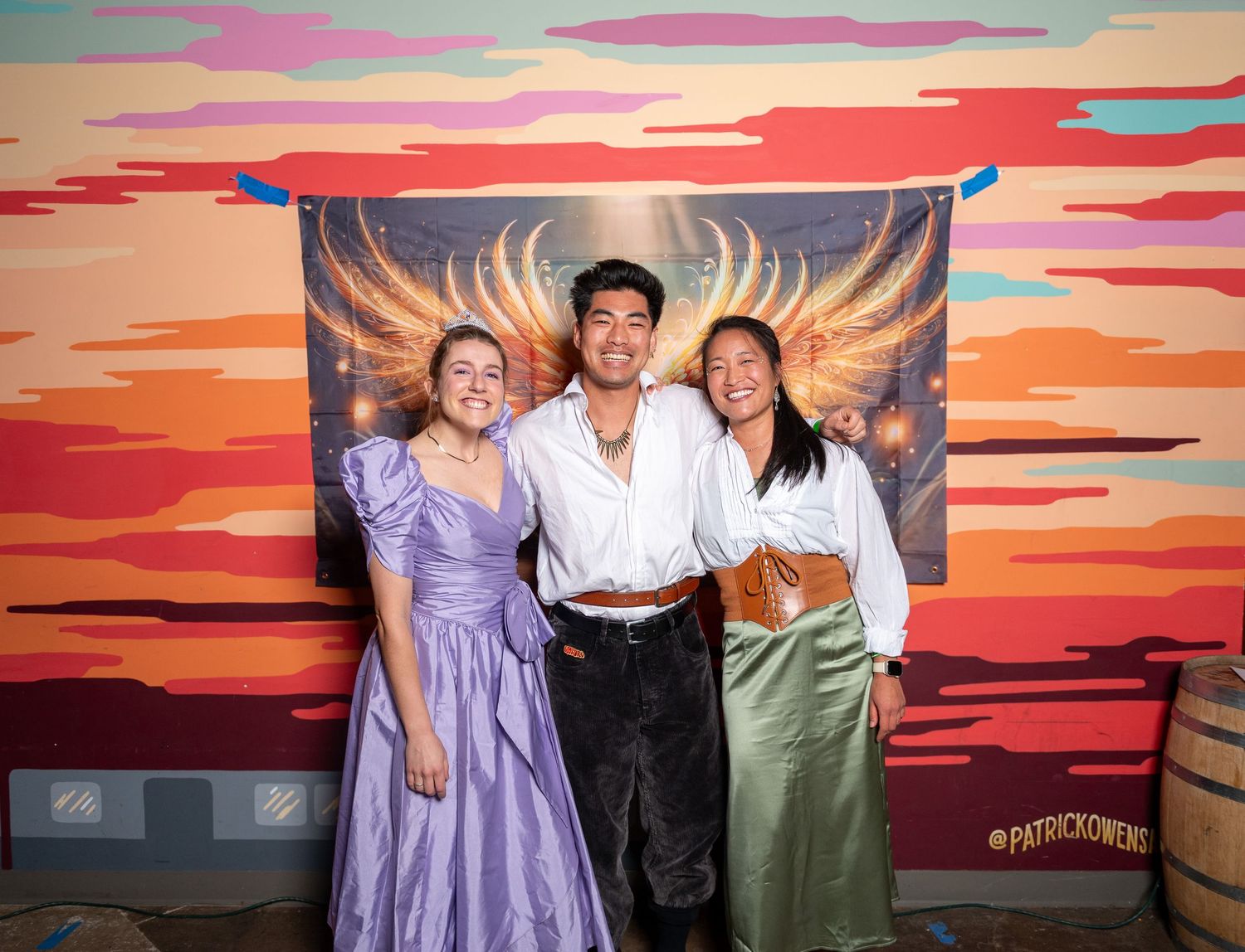 Friends gather for photos against vibrant orange and red sunset mural with angel wing artwork.
