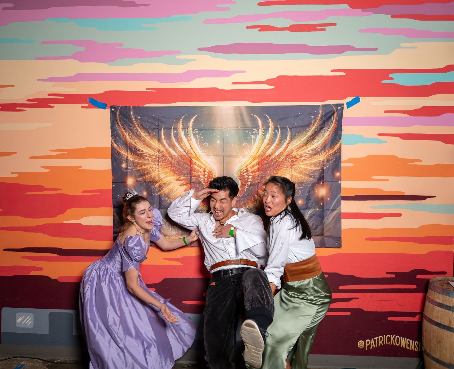 Group strikes dynamic poses against colorful sunset mural with golden wings artwork.