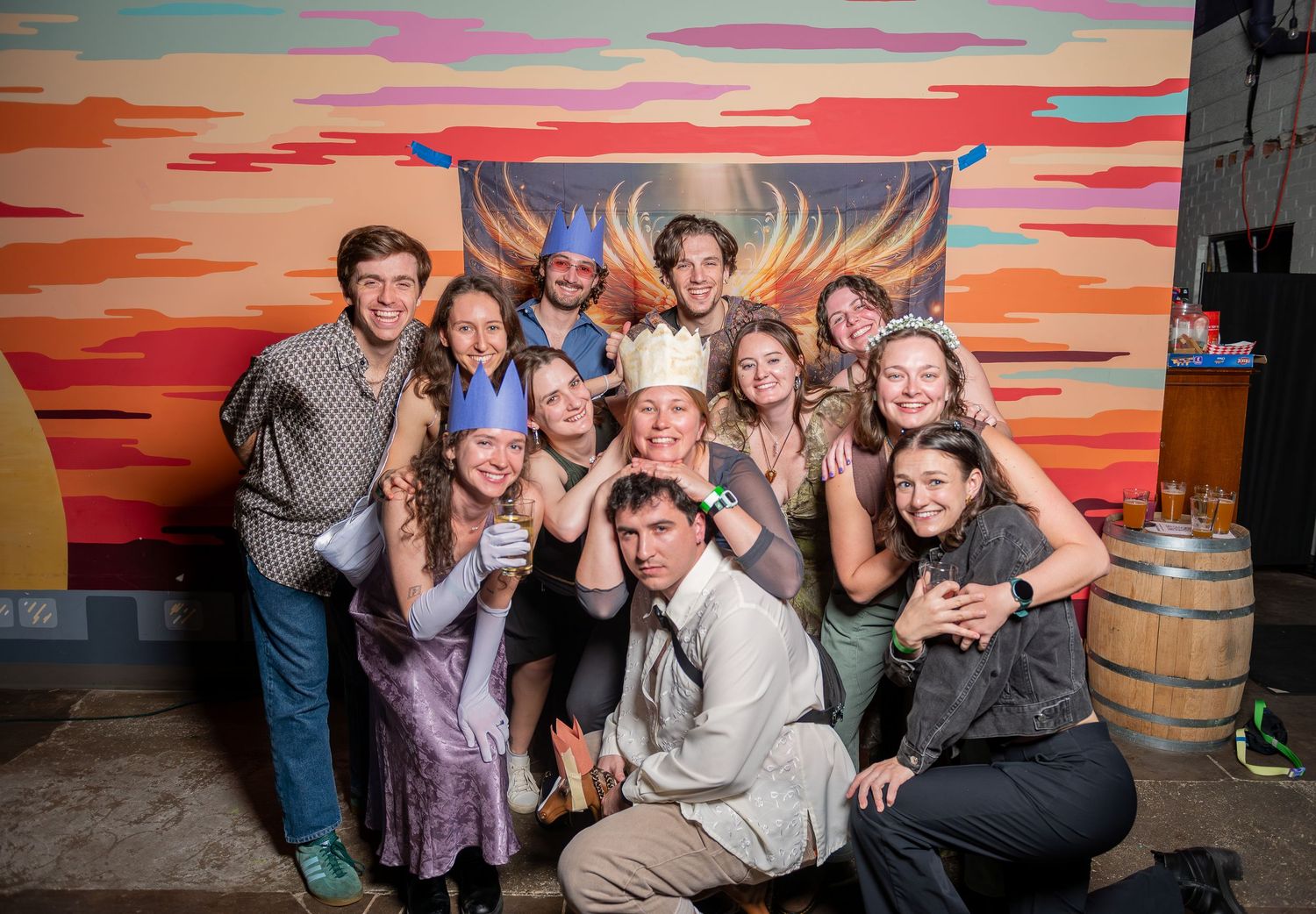 A group of people pose together against a vibrant sunset-colored wall with warm orange and pink hues.