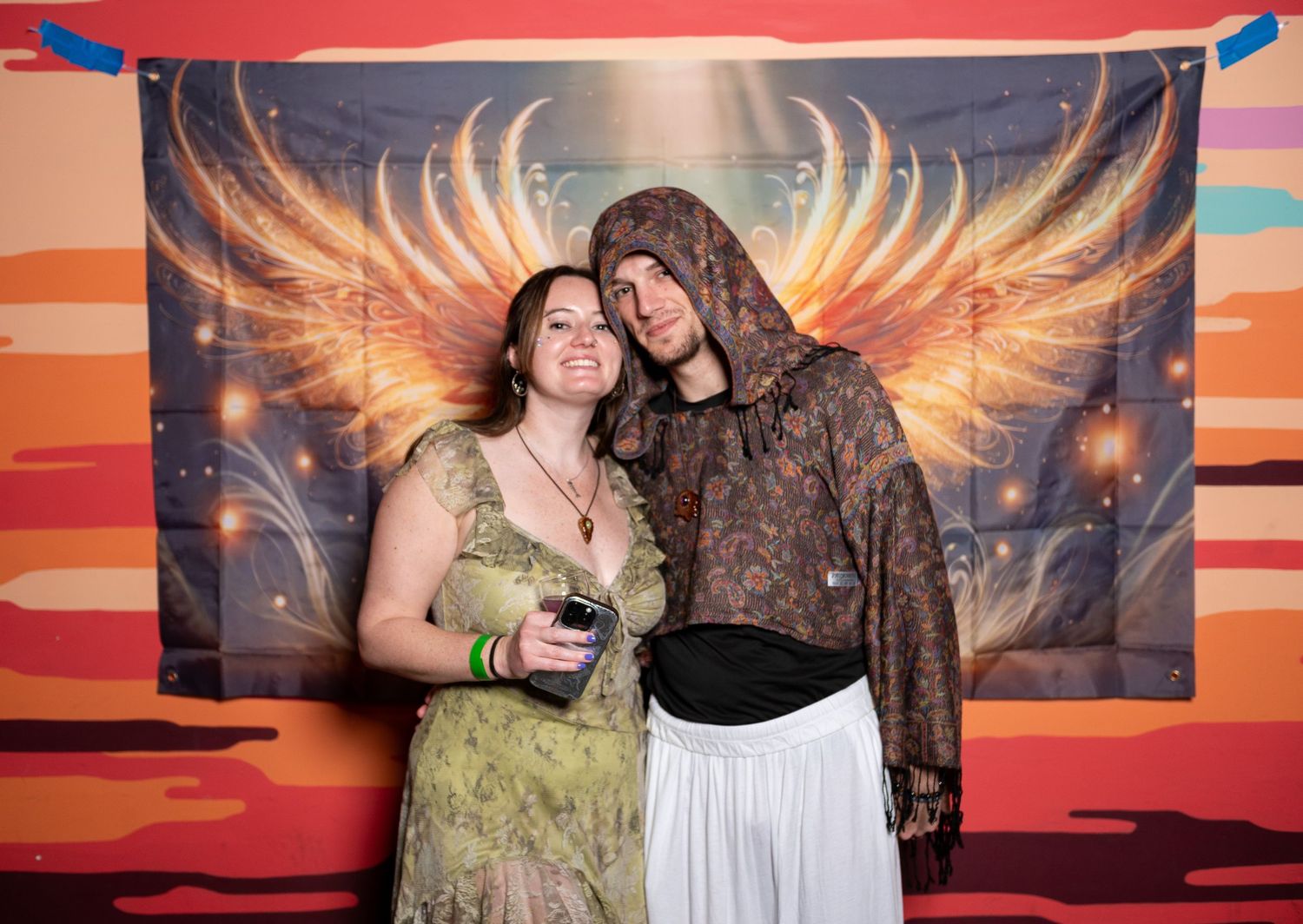 Two people pose together against a backdrop with glowing orange wings in a festive party setting.