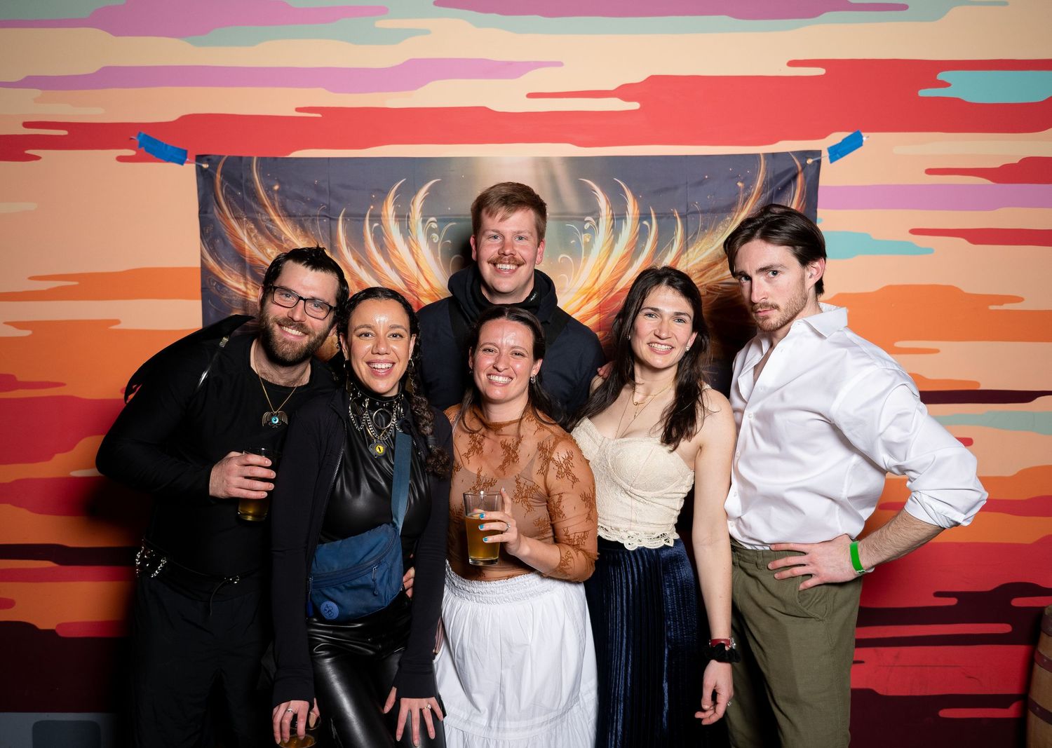Group of friends posing together at a party with vibrant orange and pink sunset-themed backdrop wall.
