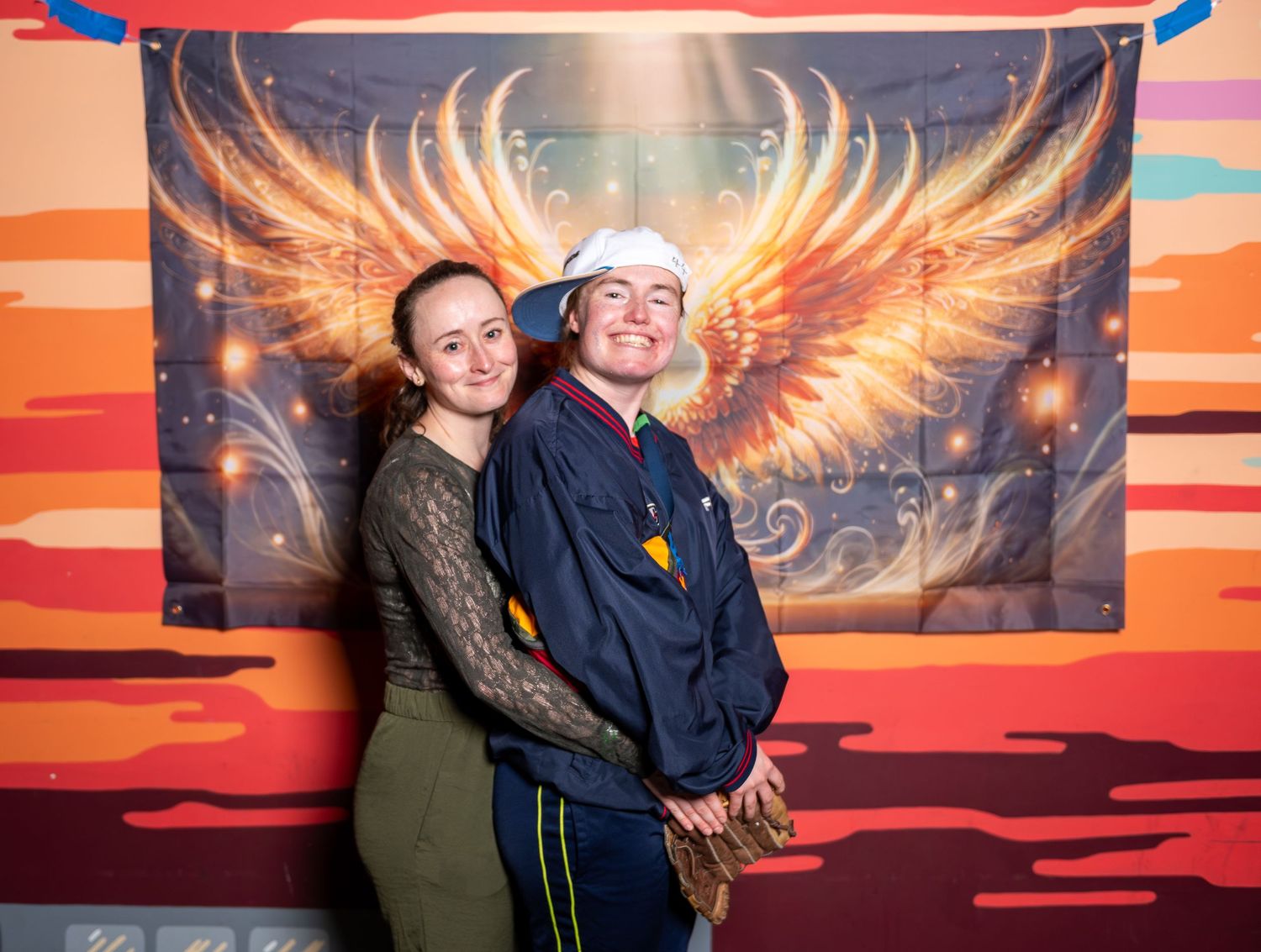 Pair poses enthusiastically in front of dramatic wing mural with orange sunset background.