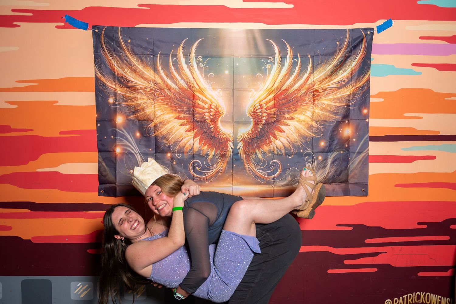 Group poses in front of vibrant sunset backdrop with glowing wings art installation at photo booth event.