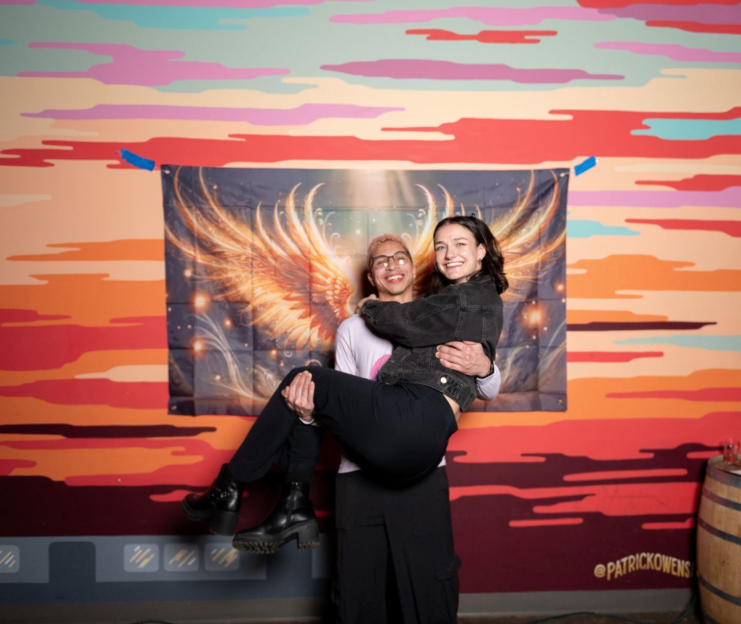 Group poses together in front of vibrant sunset-colored backdrop with glowing wings artwork.