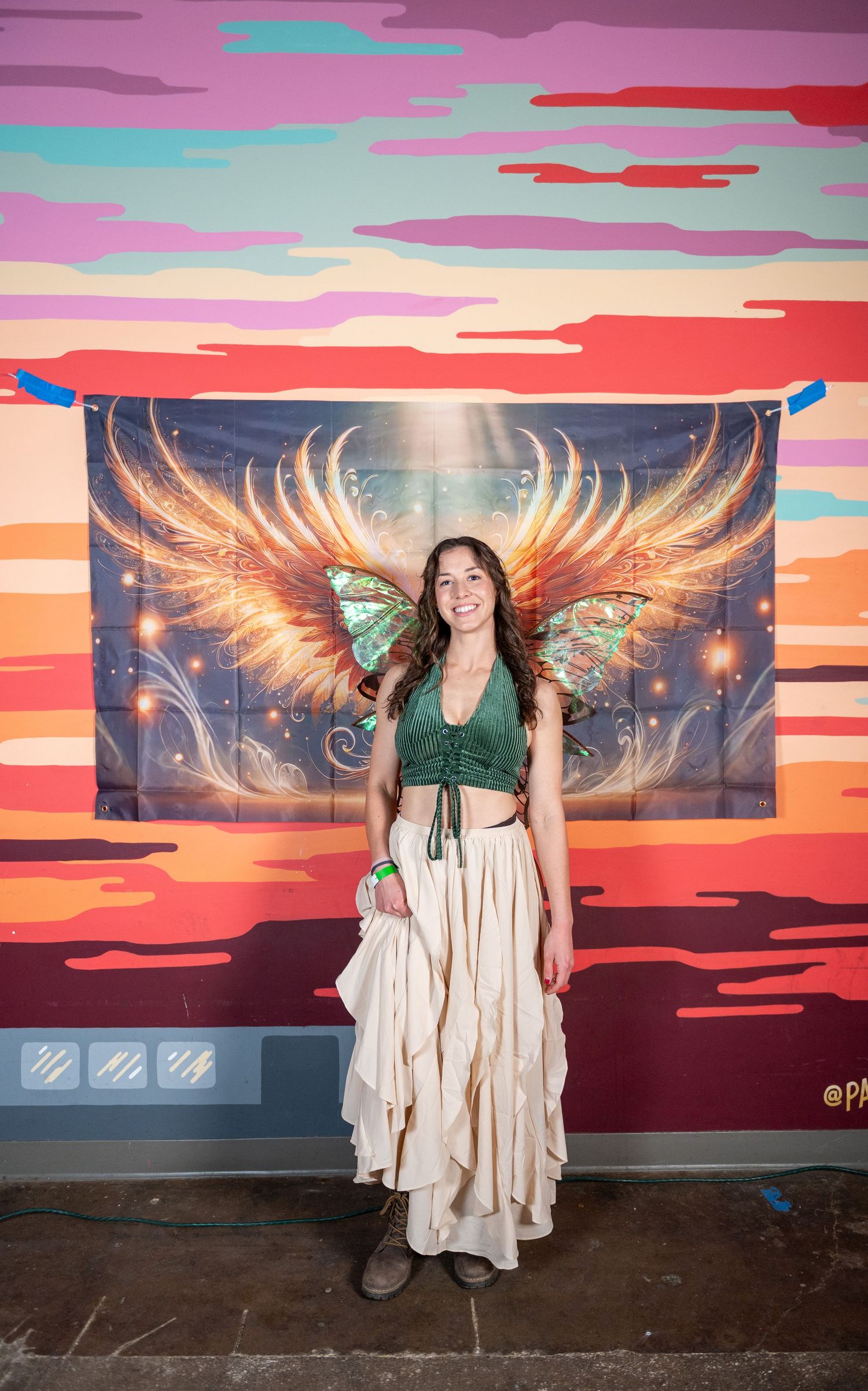 A person poses in front of a vibrant sunset mural with glowing angel wings in an urban setting.