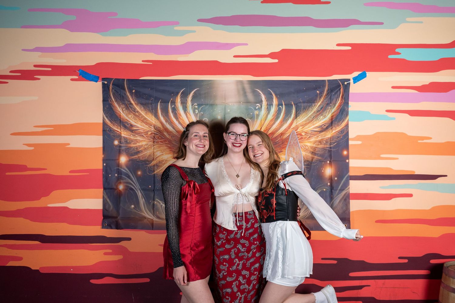 People celebrate in formal attire against dramatic sunset mural with golden wing design.