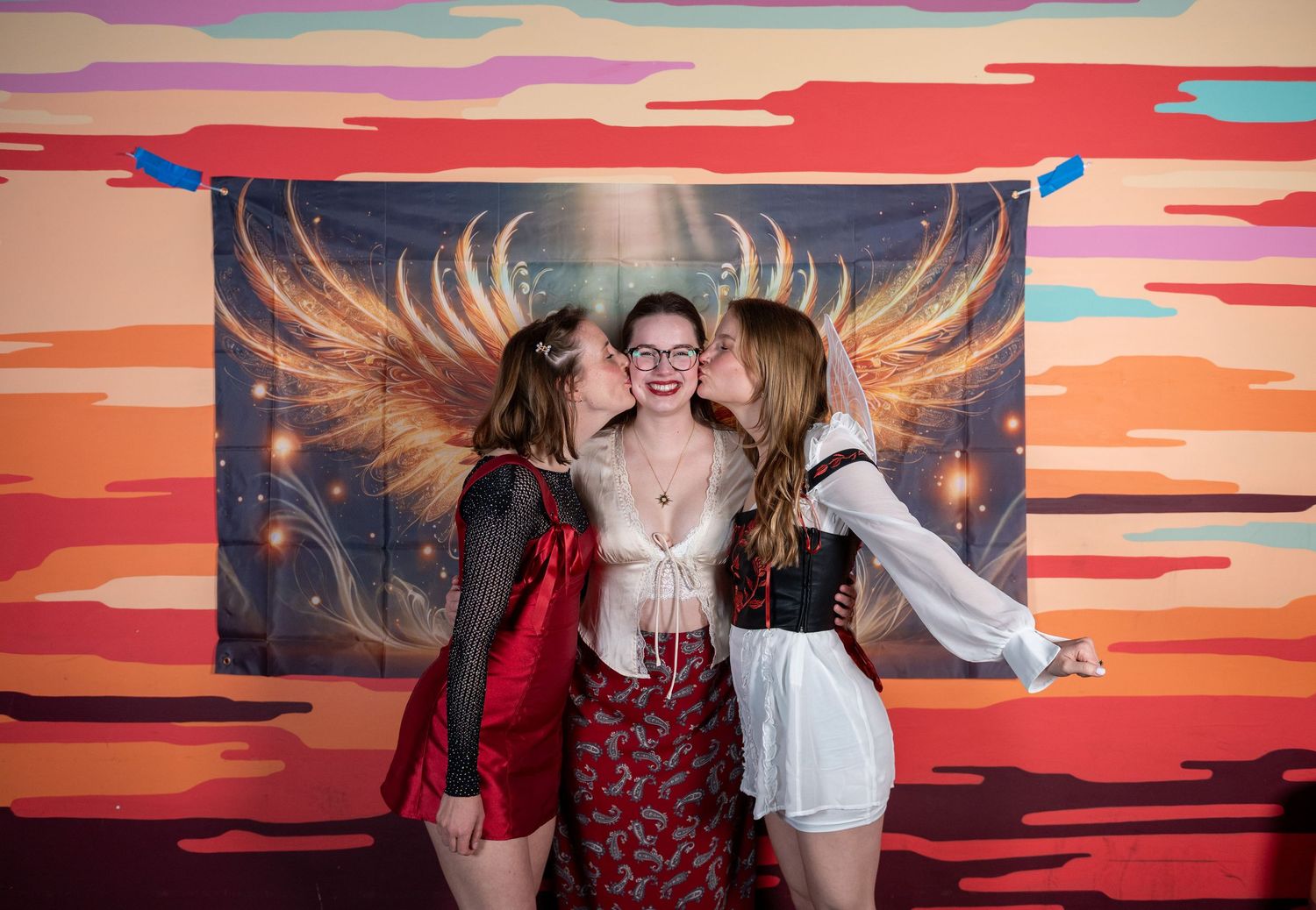 Friends pose together in front of a vibrant sunset mural with golden wings artwork on the wall.
