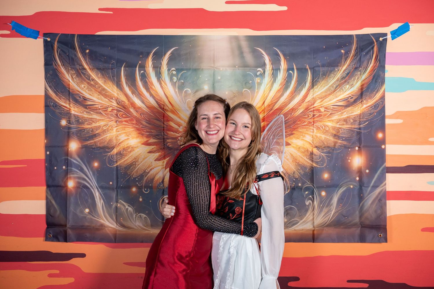 Friends gathered in front of a dramatic sunset backdrop with glowing angel wings artwork.
