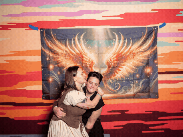 Two people embrace in a romantic pose against a dramatic sunset sky with glowing angel wings in the background.