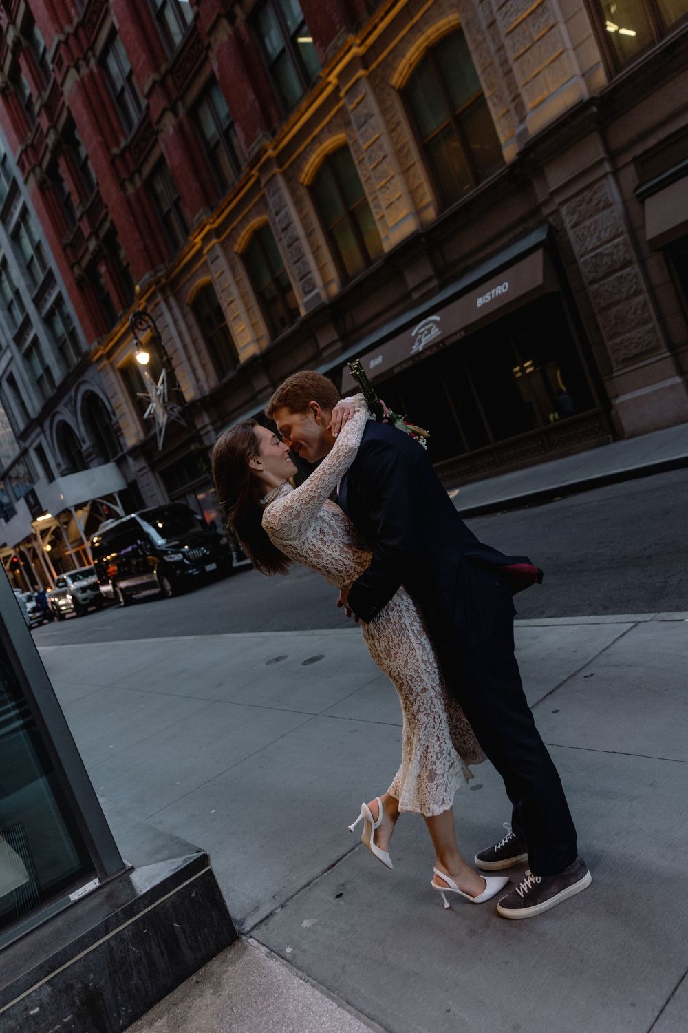 A couple dances romantically on a city sidewalk at dusk, with historic buildings in the background.