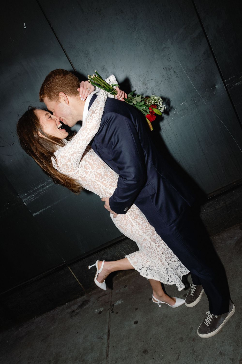 A couple in formal attire sharing a romantic dance dip pose against a dark textured wall.