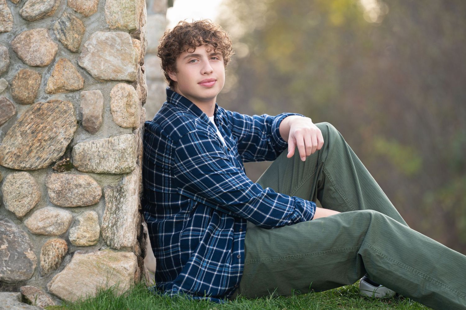 Person in blue plaid flannel shirt and white t-shirt leaning against railing outdoors with evergreen trees in background.