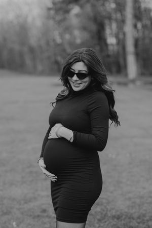 Expectant person in black dress poses outdoors in natural lighting.
