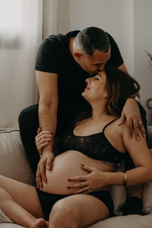 Intimate maternity photo of couple sharing tender moment on couch in black lingerie.