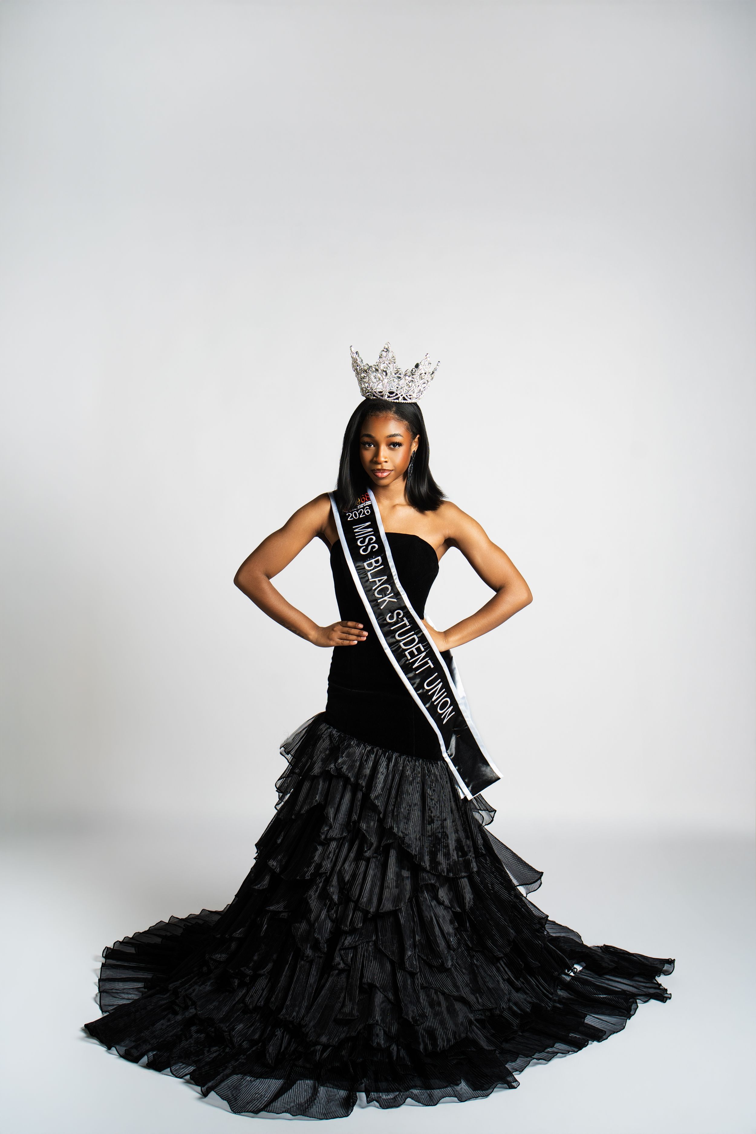 A beauty pageant winner in a dramatic black layered ball gown and crown poses against a white backdrop.