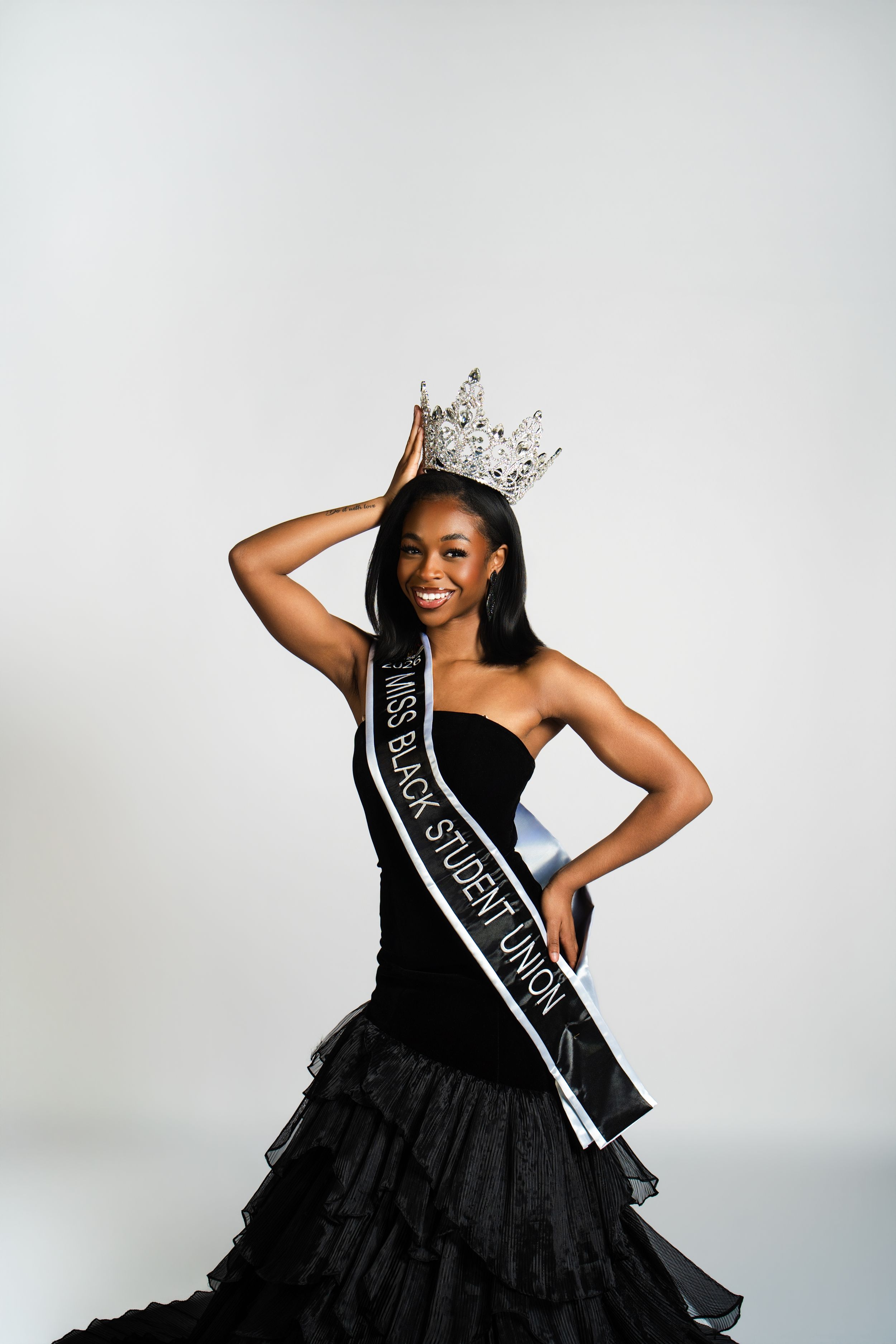 A pageant contestant in a black evening gown and crown poses for glamour shots against a light background.