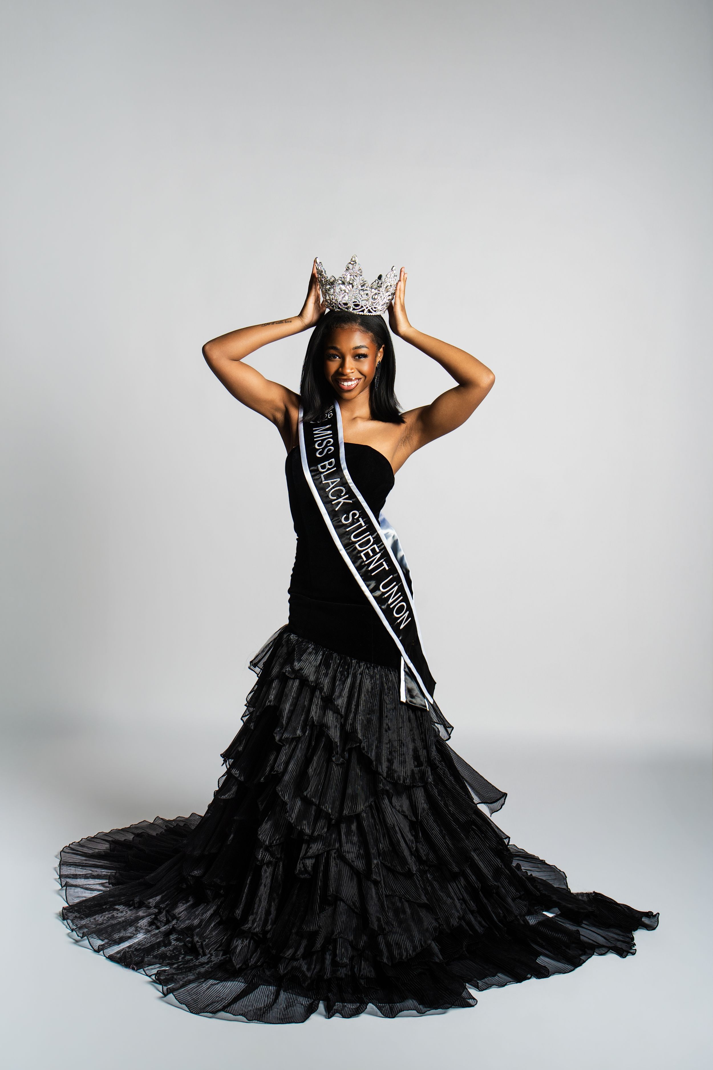 A pageant winner in a black ruffled evening gown and crown poses with hands raised on a light gray background.