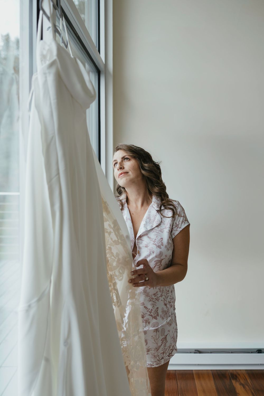 Person in white lace dress stands near window with curtains in natural light interior setting.
