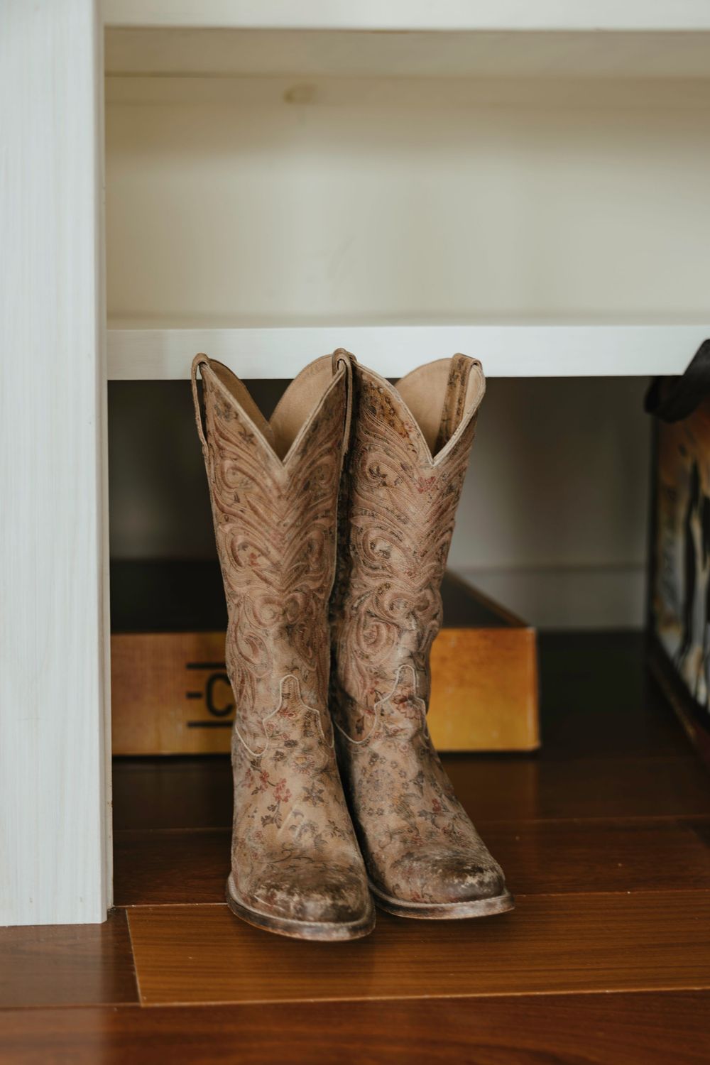 A pair of worn brown leather cowboy boots with intricate embroidery patterns sits on a wooden floor near a shelf.