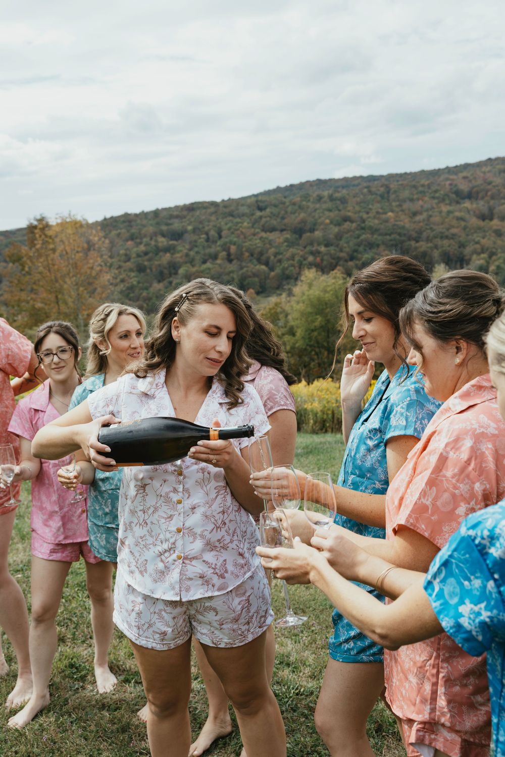 A group of people in colorful summer attire celebrate outdoors with champagne bottles on a hillside.