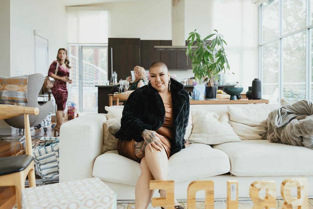 Two people sit together on a couch in a bright, modern living room with large windows and indoor plants.
