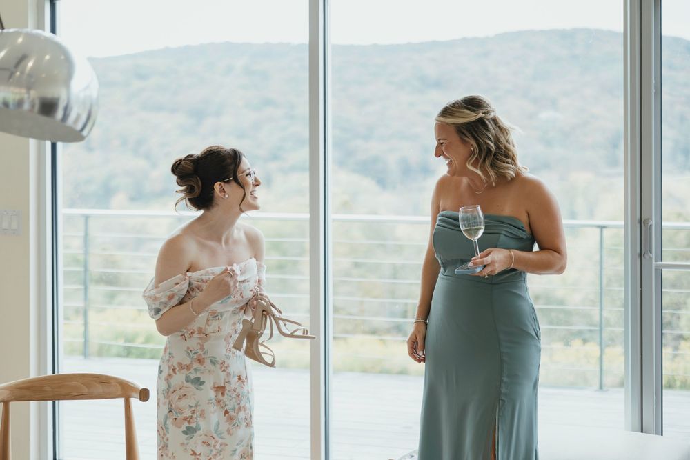 A bride and bridesmaid share an emotional embrace in front of large windows overlooking a scenic mountain view.
