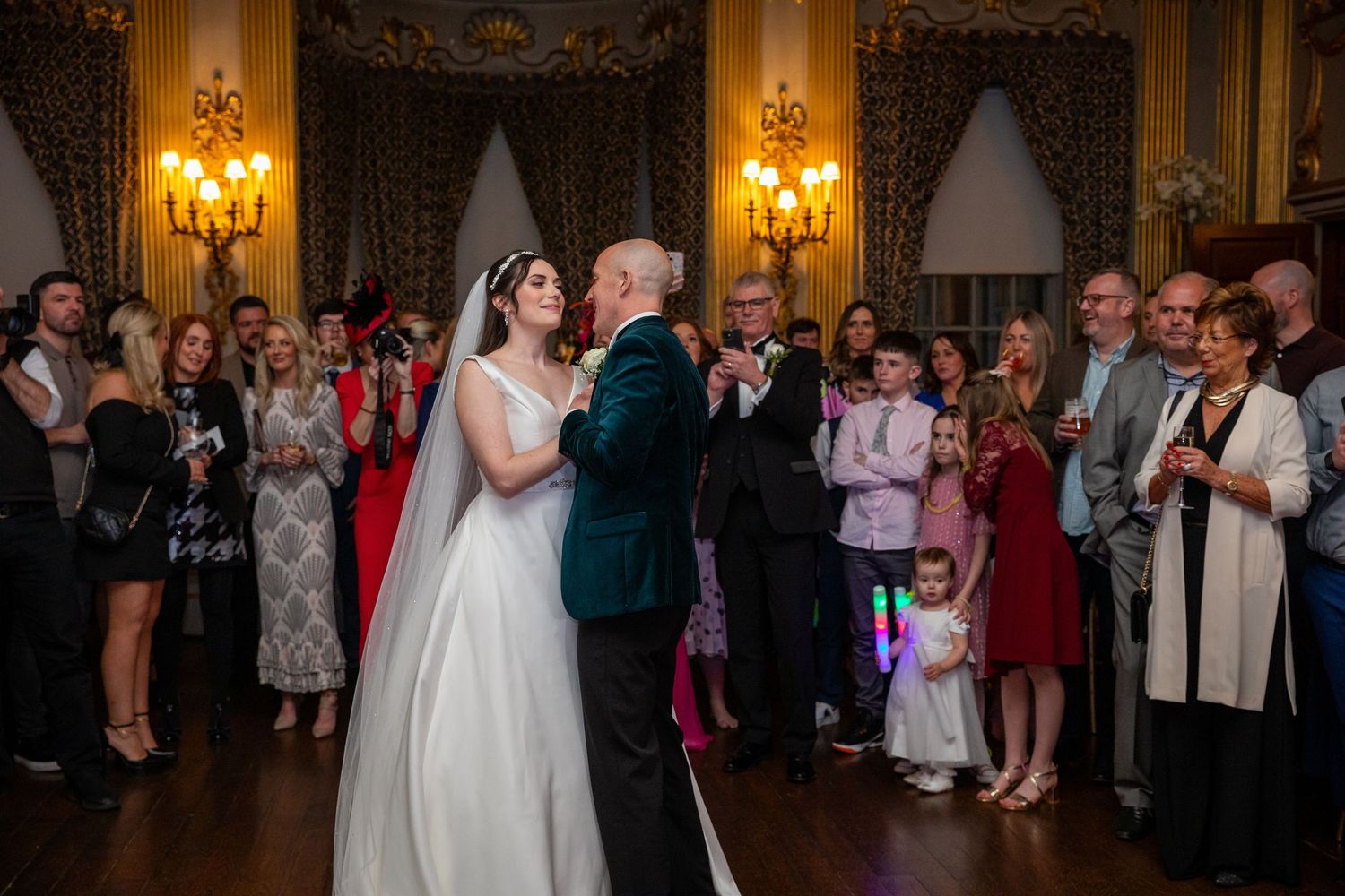 Bride and groom share their first dance surrounded by smiling wedding guests in an elegant candlelit ballroom.