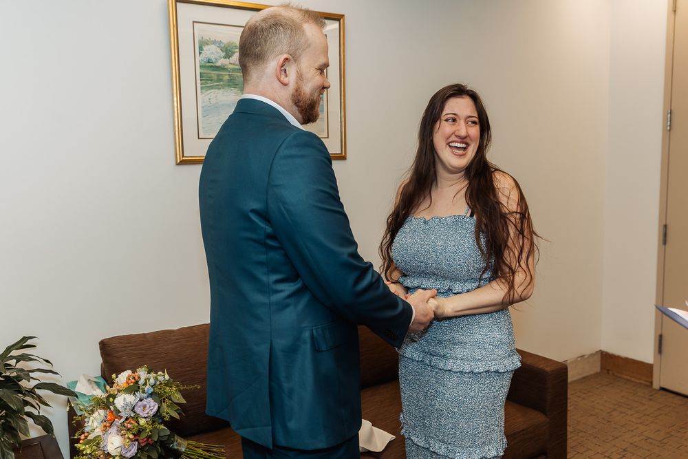 Couple holding hands and smiling in a blue suit and dress during an indoor wedding ceremony.