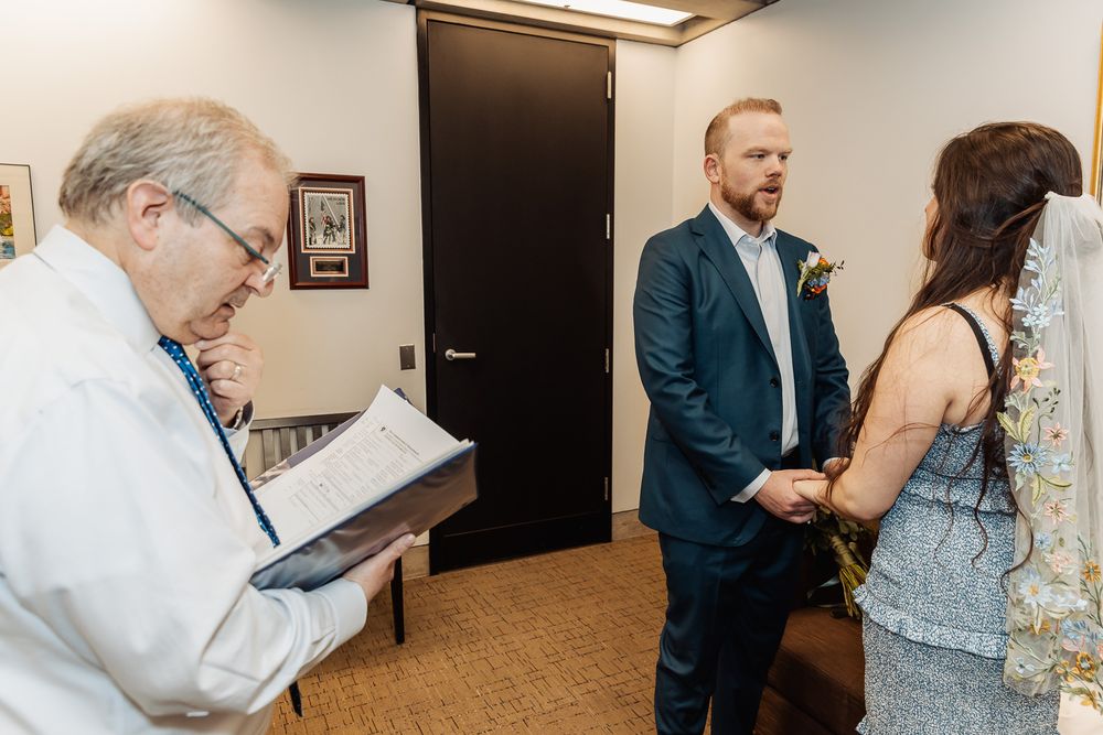 Officiant reads vows as groom in blue suit holds hands with bride during intimate indoor wedding ceremony.