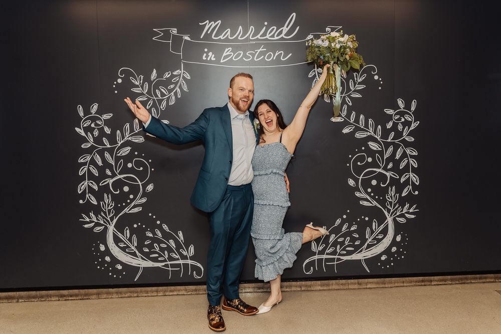 Newlyweds celebrating in front of a Married in Boston chalkboard sign with a bridal bouquet.