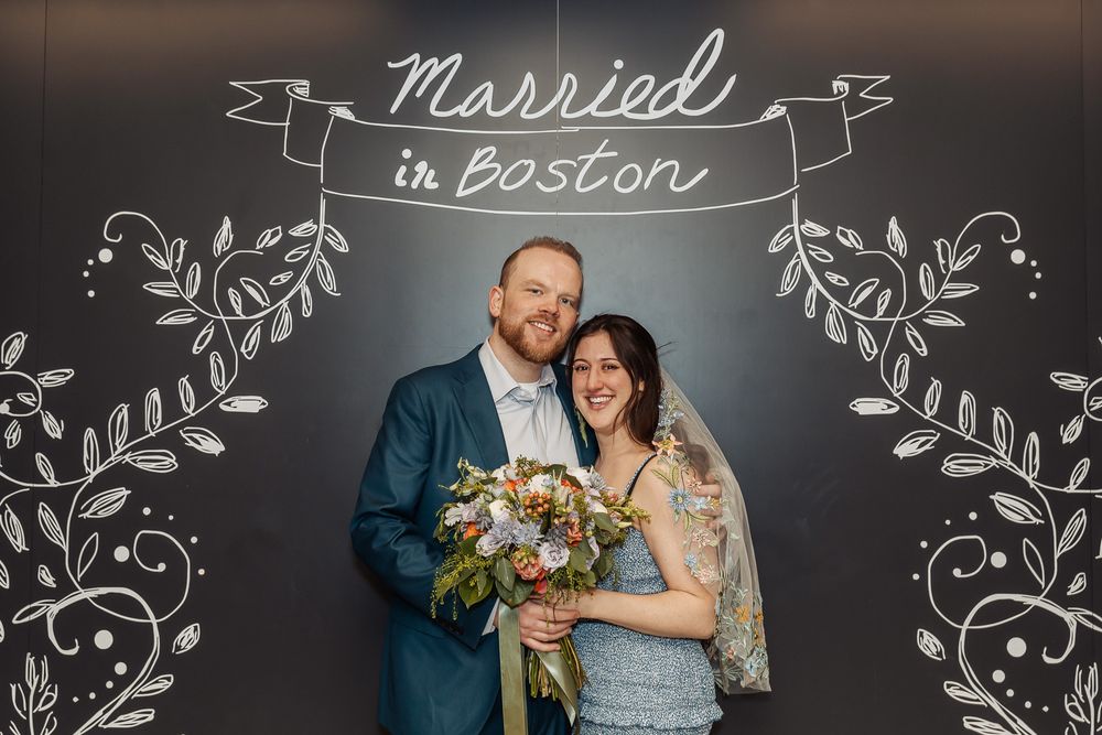 Newlyweds smiling in front of Married in Boston chalkboard sign holding bouquet.