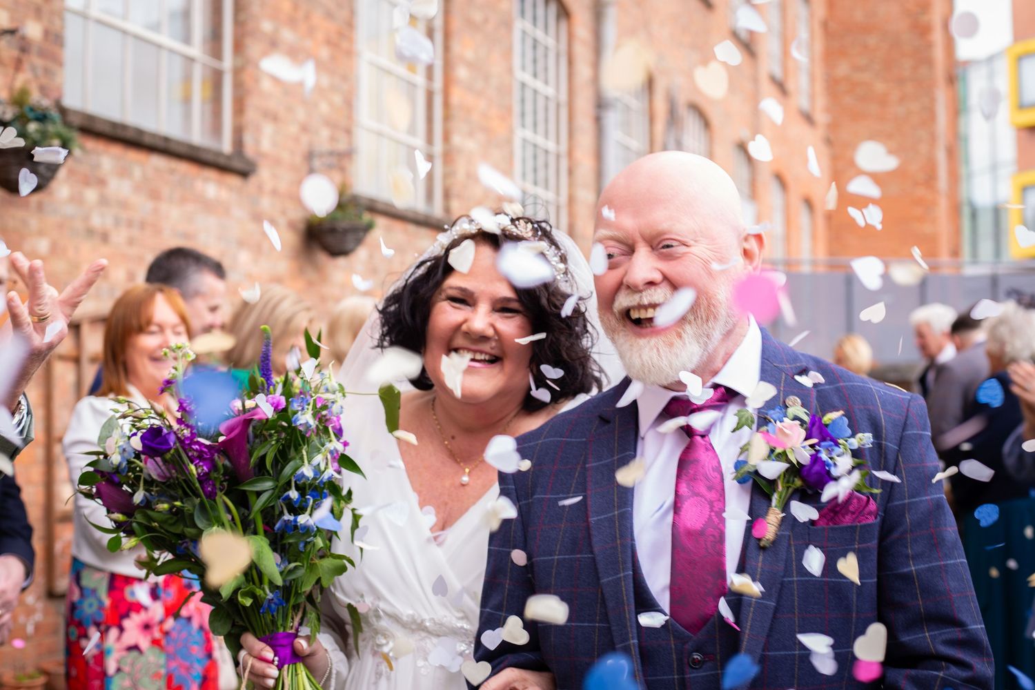 Joyful bride and groom laughing as colorful confetti falls during outdoor wedding celebration.