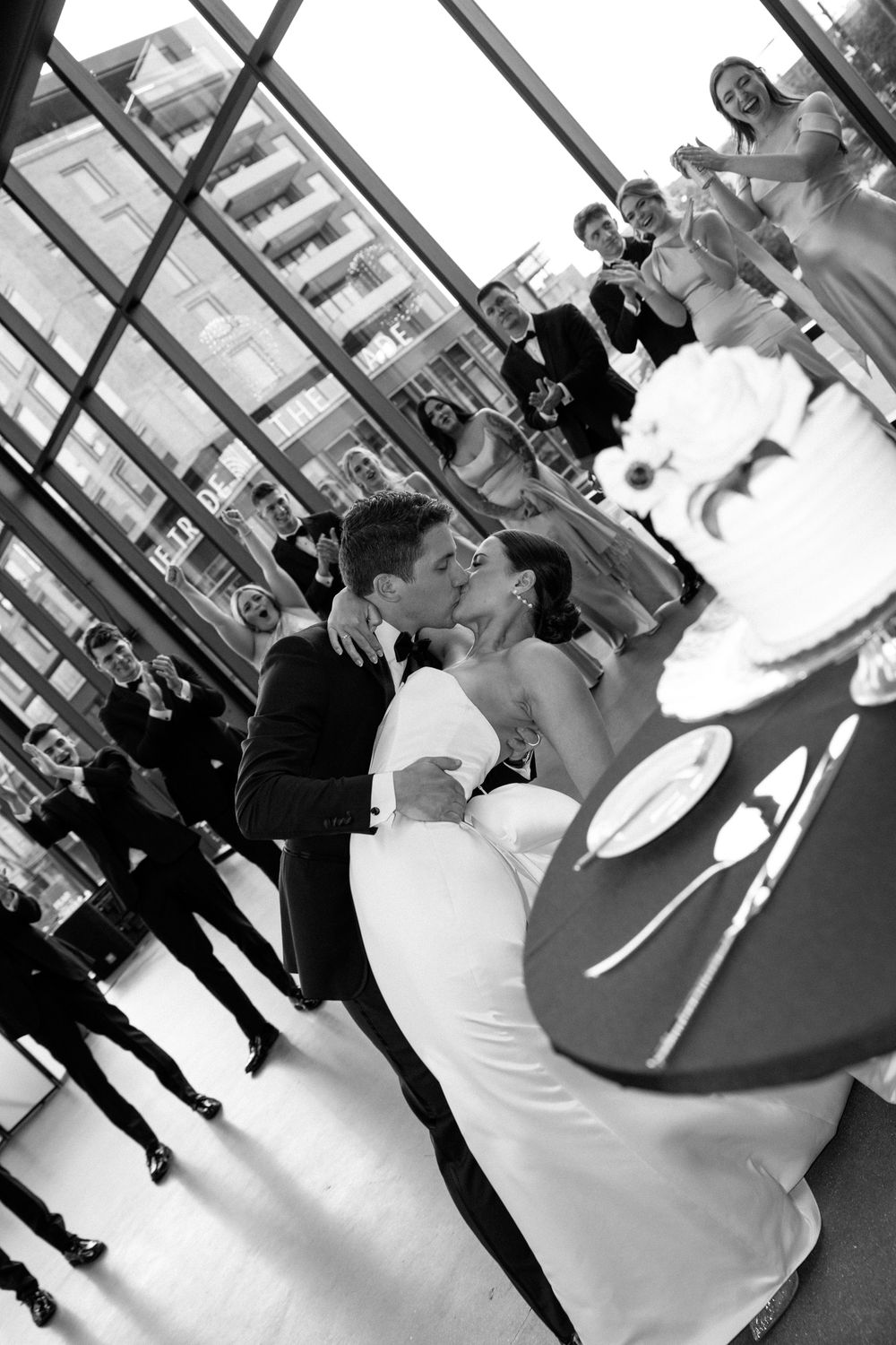 Bride and groom share a first dance kiss surrounded by wedding guests in a modern glass venue.