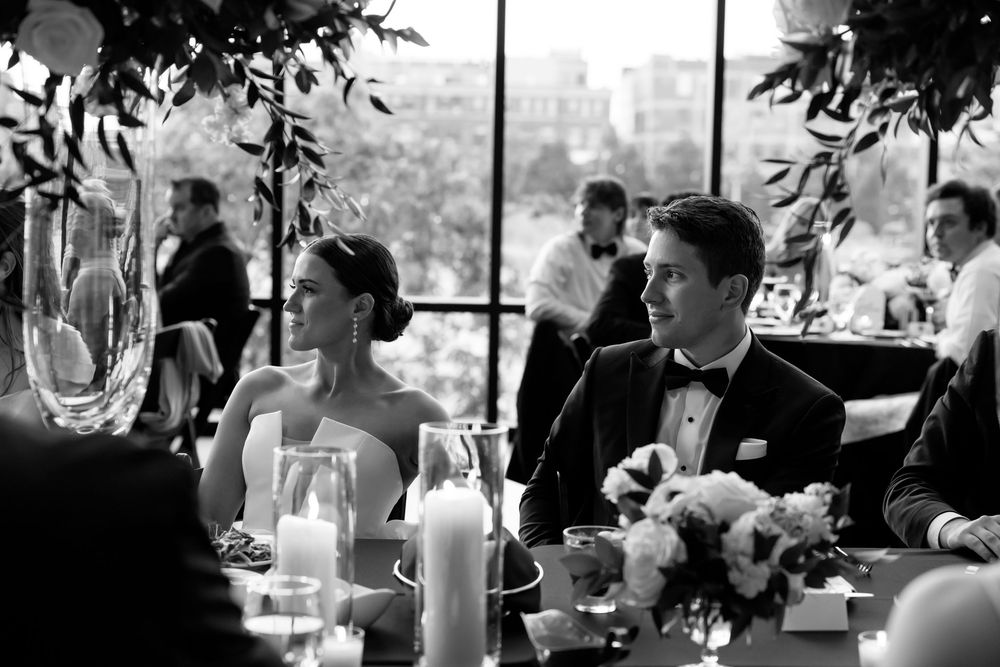 Bride and groom sit at sweetheart table during wedding reception in black and white photo.