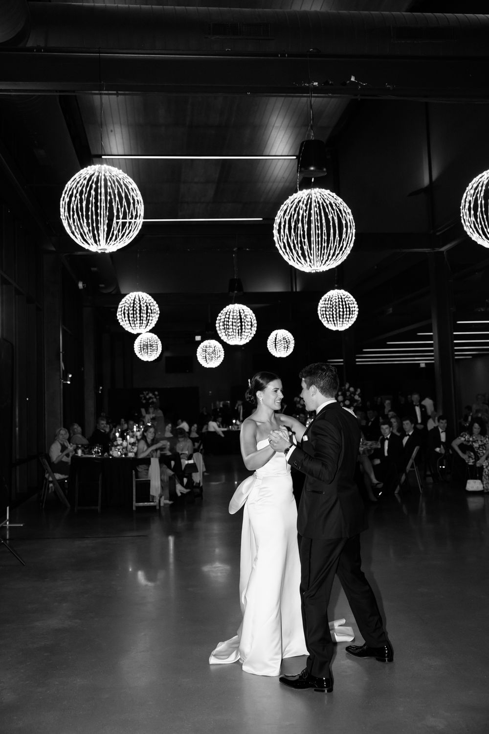 Bride and groom share first dance under glowing spherical chandeliers in elegant black and white photo.