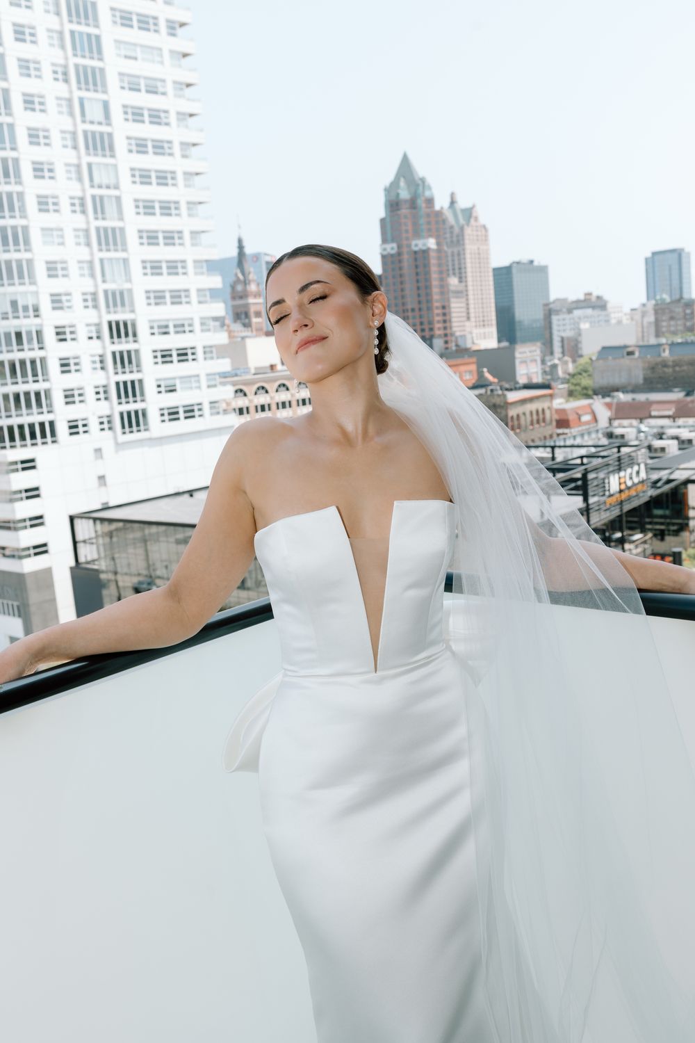Beautiful bride in strapless white gown and veil poses on rooftop balcony with urban skyline.