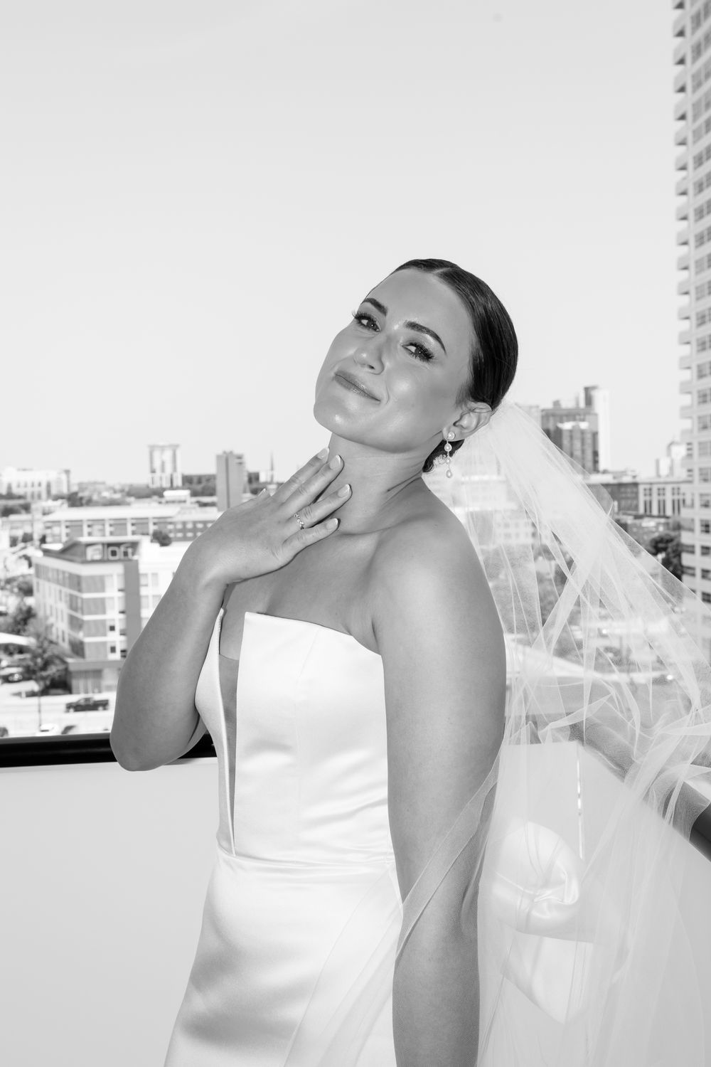A bride in a strapless white gown and veil poses gracefully on a rooftop with a city skyline behind her.