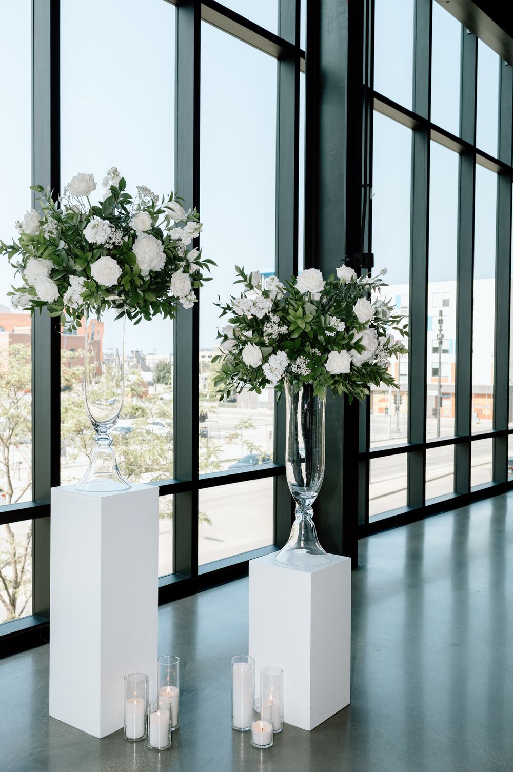 Elegant white floral arrangements on pedestals with candles in a modern venue with floor-to-ceiling windows.