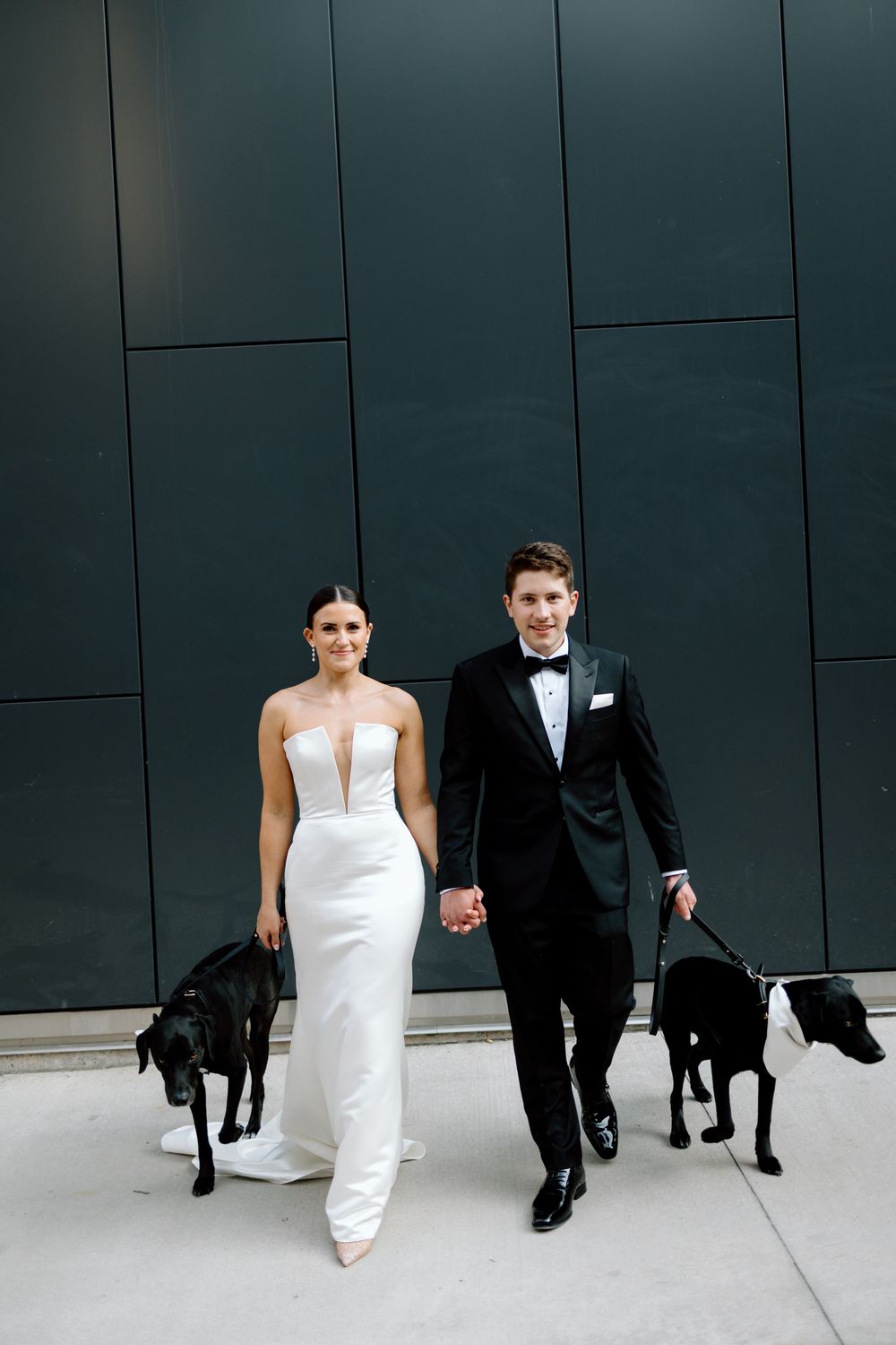 Bride in white gown and groom in black tuxedo hold hands with two black dogs in front of a dark wall.