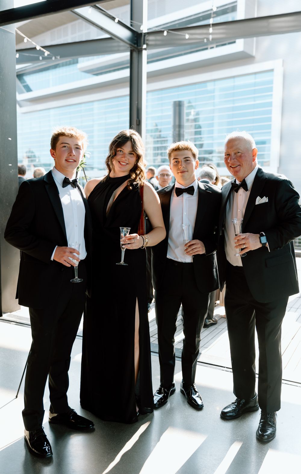 Four elegantly dressed young people in black formal attire pose together at an upscale event venue.